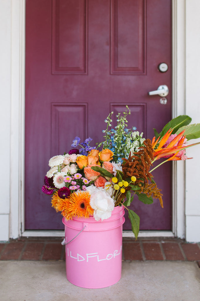 A bunch of flowers in a pink bucket in front of a maroon door. It's filled with white, orange, yellow, maroon, tan, purple, blue, and green foliage and flowers, including daisies, roses, scabiosa, billy balls, amaranthus, dahlia, delphinium, heliconia, ginger.