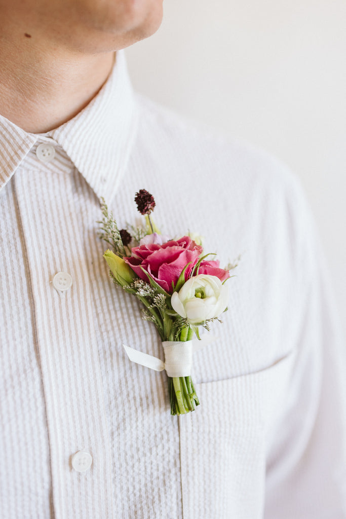 a flower boutonniere it attached to a white-striped shirt. It's made of pink, white, green, and dark purple flowers, including spray rose, thistle, ranunculus, and other foliage, collected with a white ribbon.