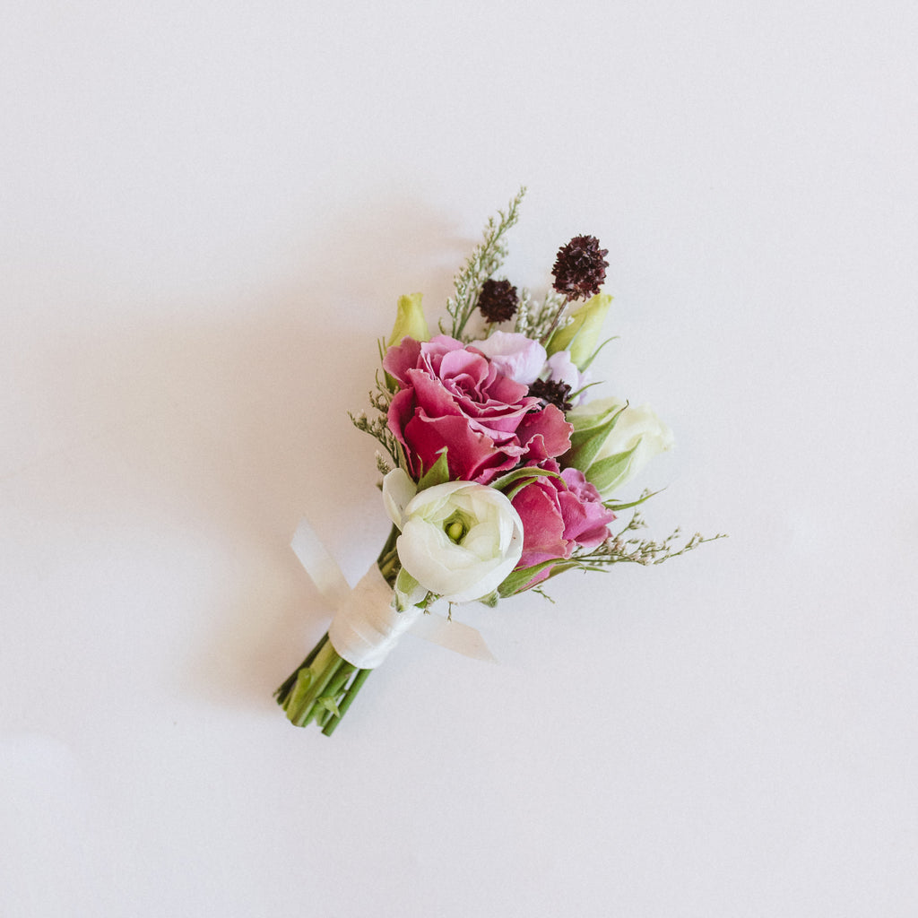 A flower boutonniere made of pink, white, green, and dark purple flowers, including spray rose, thistle, ranunculus, and other foliage, collected with a white ribbon on a white background.