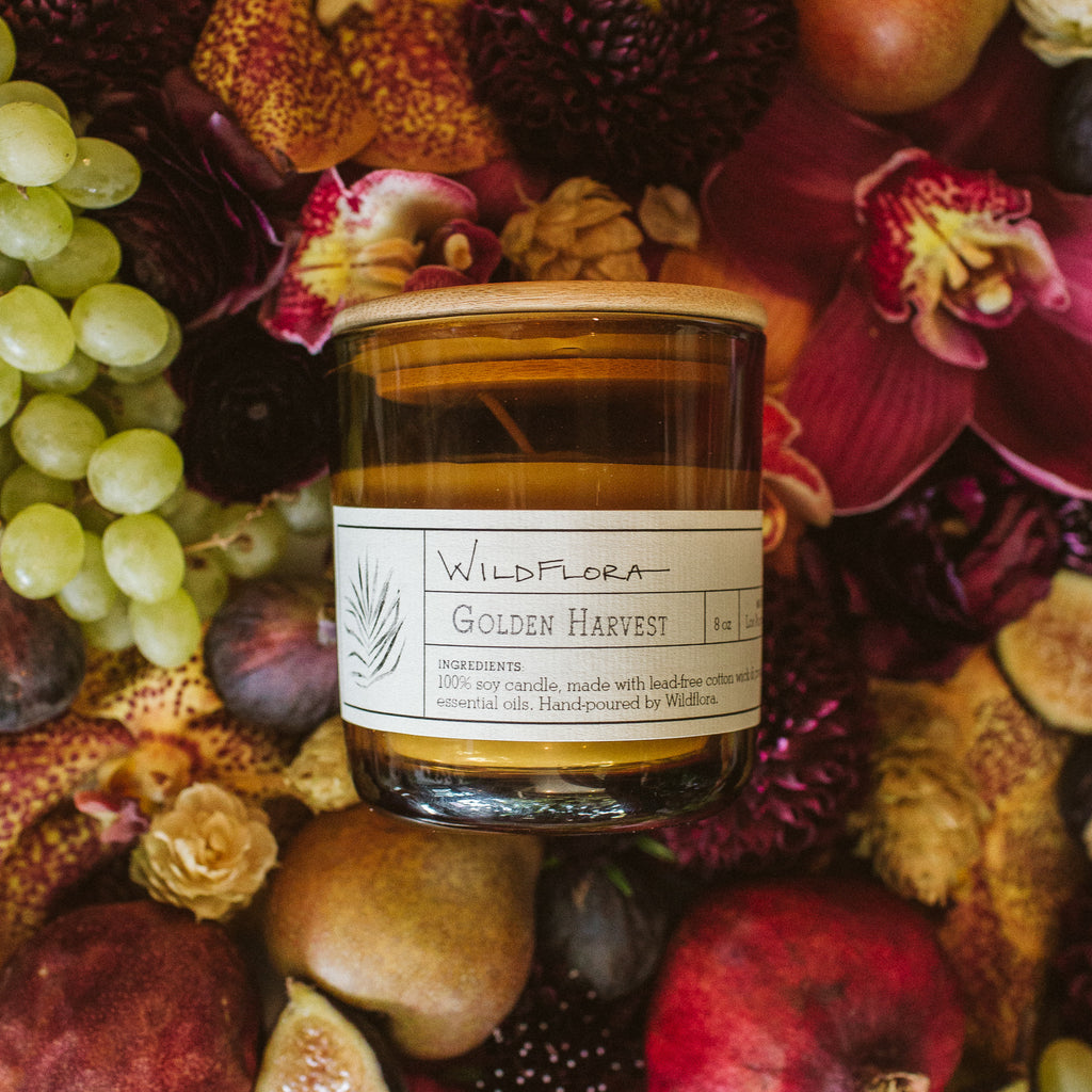 A single-wick WildFlora candle with a label reading 'Golden Harvest' placed among various fruits and flowers.