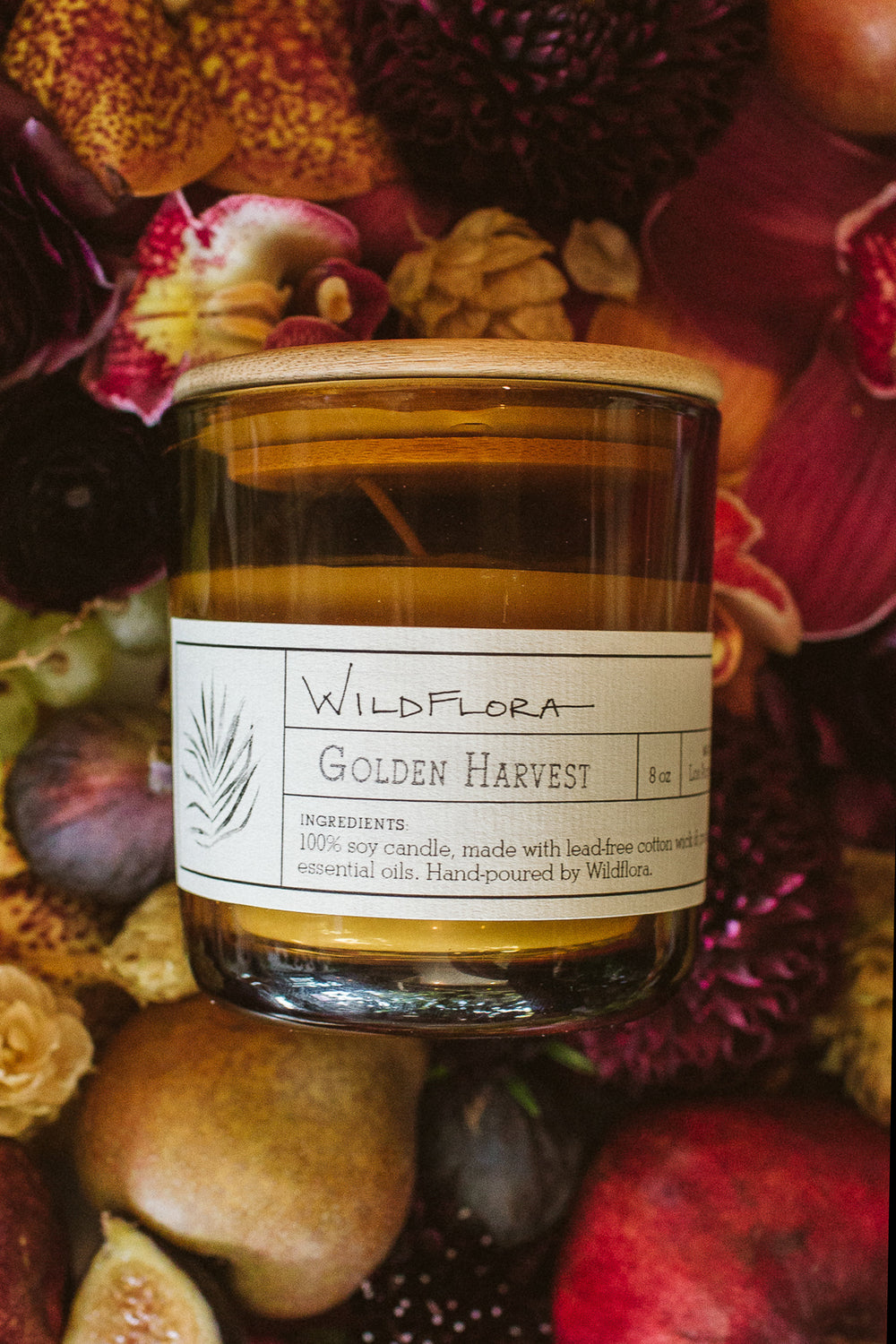 A single-wick WildFlora candle with a label reading 'Golden Harvest' placed among various fruits and flowers.
