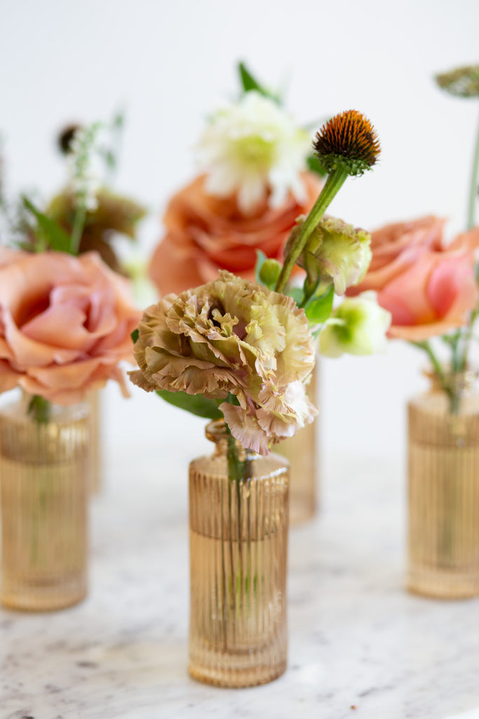 WildFlora's Gathered Garden Bud Vases in tan ribbed glass vases a on marble table. They include dusty coral-orange, blush, brown, and tan earth-toned flowers, complete with Moab roses, Queen Anne's lace, lisianthus, echinacea, and veronica.