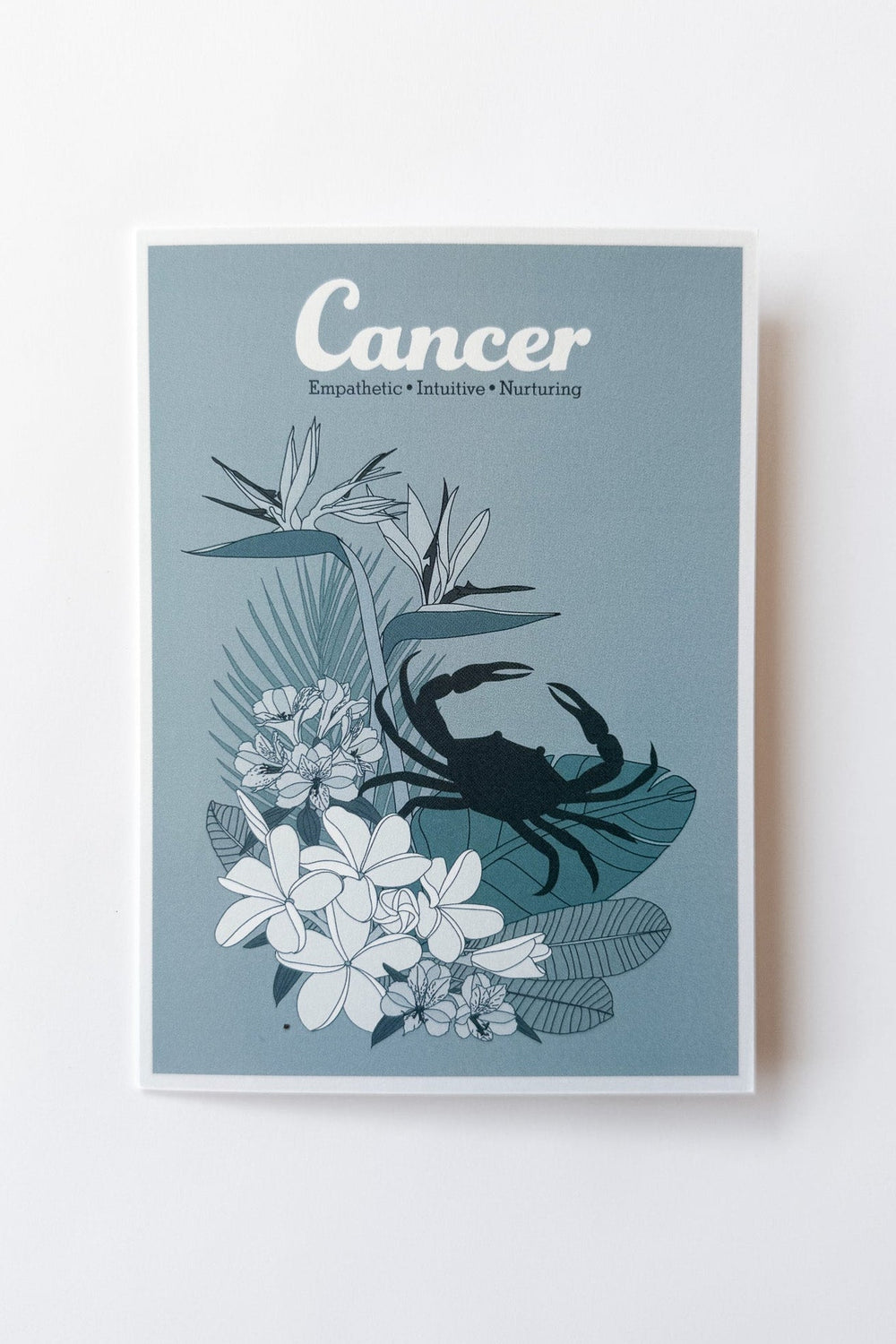 A vertical greeting card featuring a Cancer zodiac design with a blue crab silhouette over white tropical flowers and leaves on a muted blue background, shown against a plain white background.