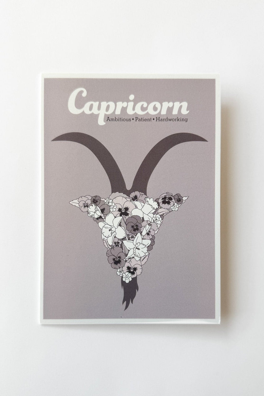 A greeting card featuring a stylized Capricorn illustration with a horned goat head made of white and violet flowers on a muted purple background, with the word 'Capricorn' printed at the top.