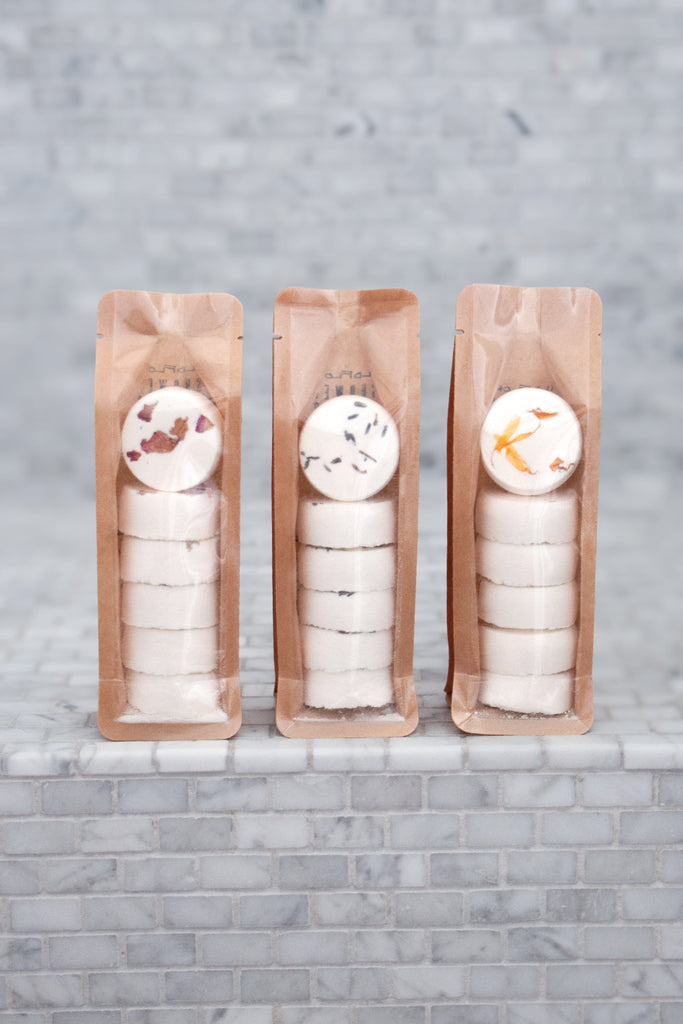 Three packets of WildFlora shower steamers with various floral designs on them, placed on a tiled surface.