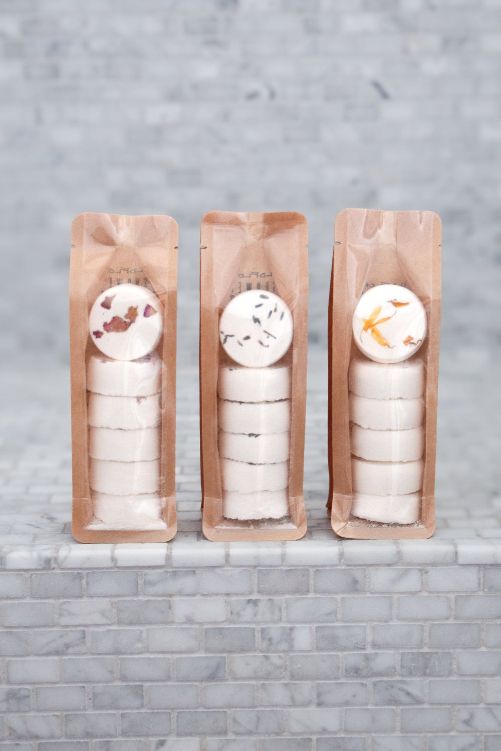 Three packets of WildFlora shower steamers with various floral designs on them, placed on a tiled surface.