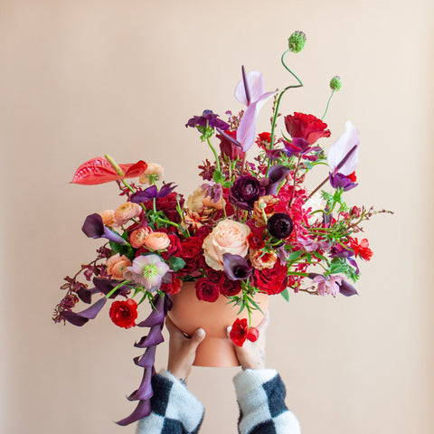 A woman in a checkered sweater holds a large flower arrangement in a terracotta container in front of a tan backdrop. It's filled with red, purple, peach, maroon, and dark violet-colored flowers, including rose, ranunculus, orchid, calla lily, anthurium, hellebore, and allium. 