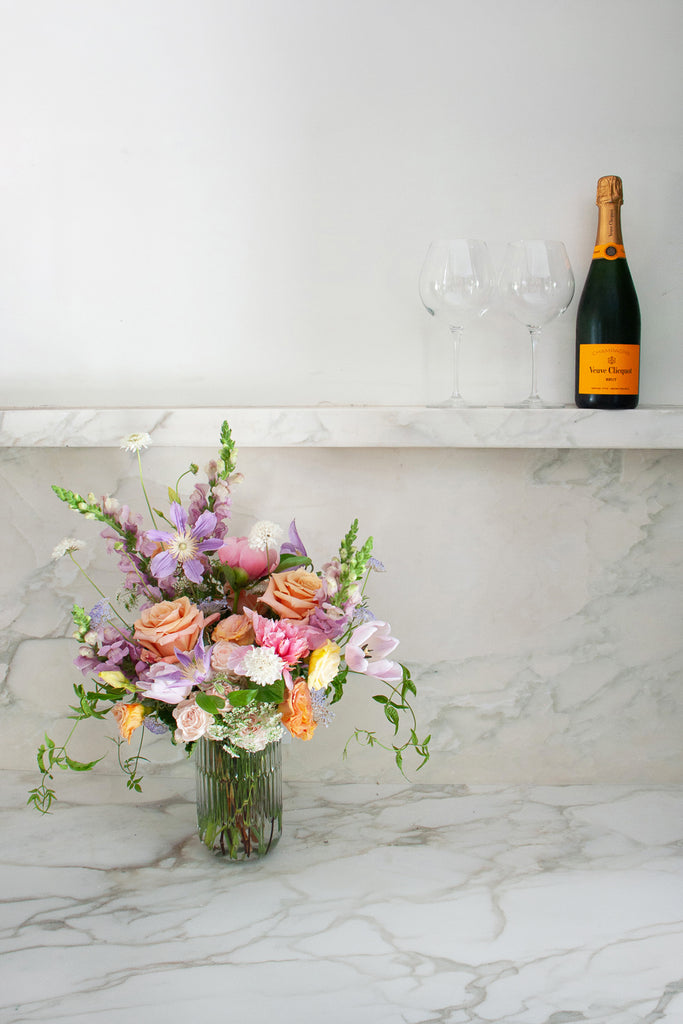 A coral, peach, pink, purple, lavender, and white flower arrangement on a marble bar with a glass of wine and two wine classes in the back for scale. The tall glass vase includes roses, tulips, clematis, allium, and snapdragon, and queen anne's lace. The vibe is sweet, romantic, and dreamy.