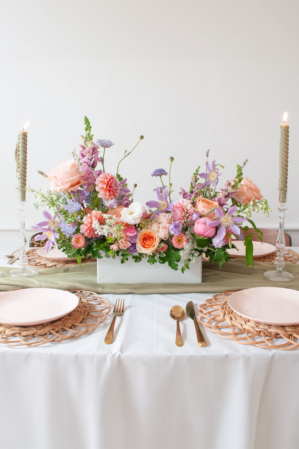 An orange, peach, pink, purple, lavender, chartreuse and white long & low flower centerpiece sits on an event table with pink plate, gold cutlery, woven plate chargers, and tall candlesticks. The floral arrangement includes roses, dahlias, clematis, peonies, allium, snapdragon, queen anne's lace, delphinium, and lisianthus. The vibe is sweet, romantic, and dreamy.