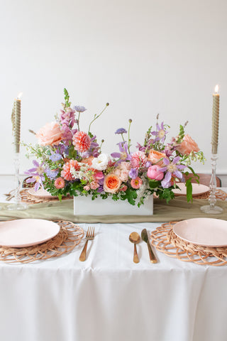 An orange, peach, pink, purple, lavender, chartreuse and white long & low flower centerpiece sits on an event table with pink plate, gold cutlery, woven plate chargers, and tall candlesticks. The floral arrangement includes roses, dahlias, clematis, peonies, allium, snapdragon, queen anne's lace, delphinium, and lisianthus. The vibe is sweet, romantic, and dreamy.
