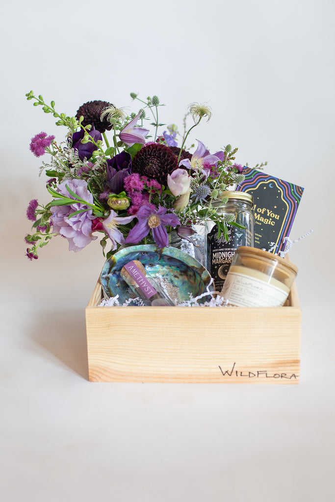 A Wildflora gift box with flower arrangement in shades of purple, abalone shell, amethyst stones, candle, drink mix, and magic card deck in a wood box.