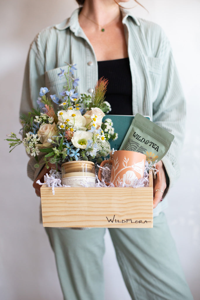 Woman holding a WildFlora gift box with white, peach, and blue flowers, a mug, loose leaf tea, and a journal in a wooden box.