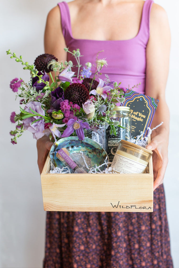 Woman holding a Wildflora gift box with flower arrangement in shades of purple, abalone shell, amethyst stones, candle, drink mix, and magic card deck in a wood box.