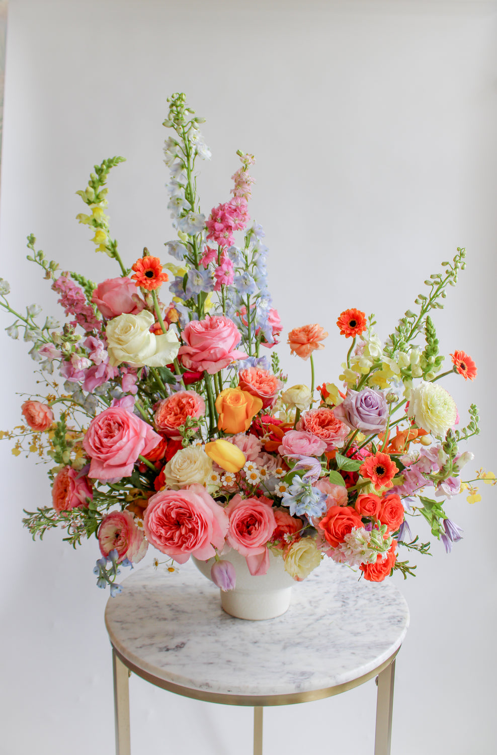 A flower arrangement in a glazed white compote vase in front of a white backdrop on a marble table. It's filled with pink, yellow, peach, orange, blue, purple, and white-colored flowers, including rose, ranunculus,, piccolini daisy, tulip, oncidium orchid, chamomile, snapdragon, and delphinium. The vibes are very Bridgerton.