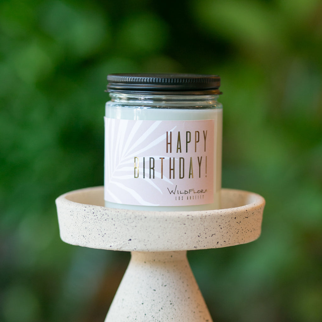 Expressions candle that says "happy birthday!" sits on a candle holder in front of greens. Single-wick, white soy wax candle in a glass container with a black lid.