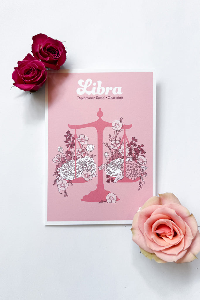 A Libra-themed greeting card featuring a pink background with a pink balance scale decorated with white and dark pink flowers, and the word 'Libra' printed at the top, shown against a white background with 3 roses.