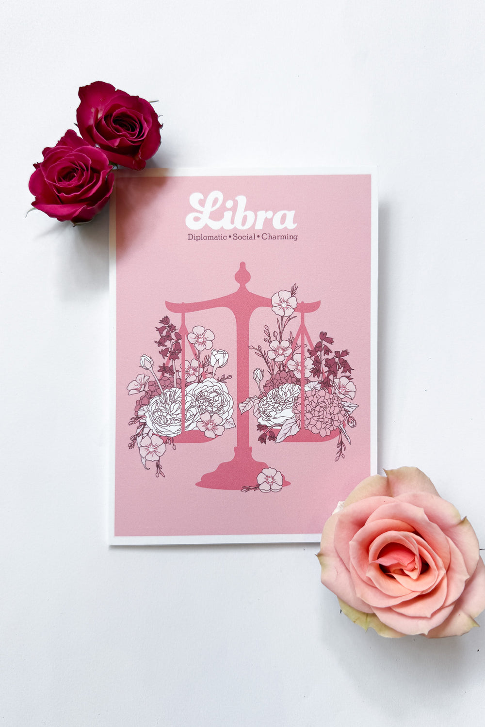 A Libra-themed greeting card featuring a pink background with a pink balance scale decorated with white and dark pink flowers, and the word 'Libra' printed at the top, shown against a white background with 3 roses.