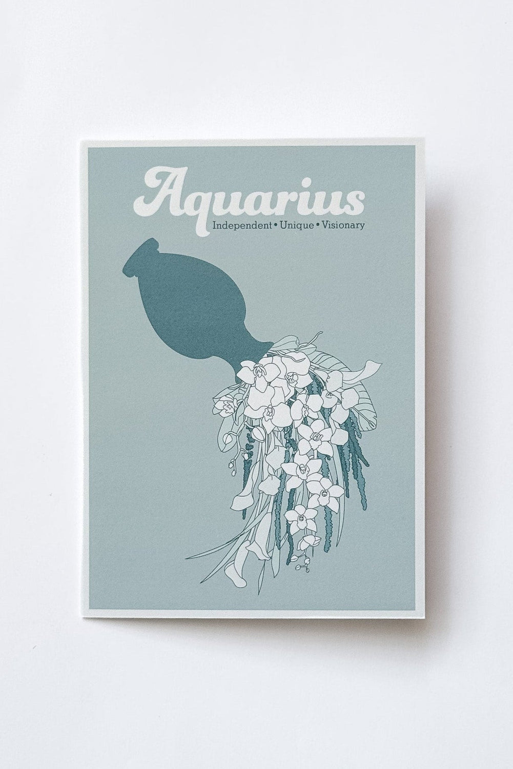 A greeting card featuring the word 'Aquarius' in white script above a teal silhouette of a jug pouring illustrated florals on a muted blue-green background, photographed against a white background.