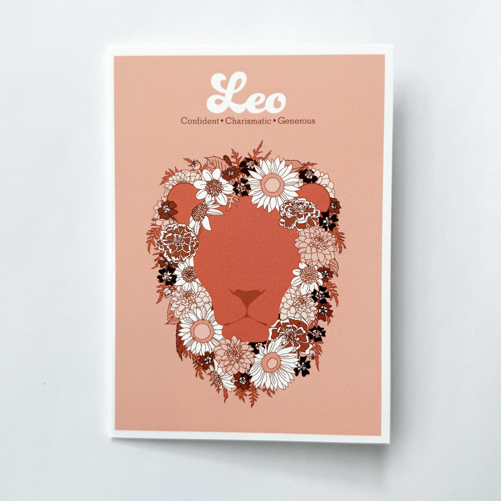 A greeting card featuring a stylized lion face silhouette in terracotta, framed by a floral mane in white, pink, and dark brown. The print has a peach background with the word 'Leo' and adjectives printed in white at the top, and is photographed against a white background.