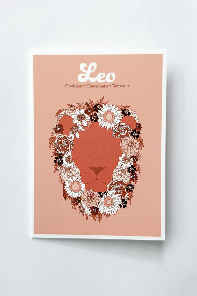 A greeting card featuring a stylized lion face silhouette in terracotta, framed by a floral mane in white, pink, and dark brown. The print has a peach background with the word 'Leo' and adjectives printed in white at the top, and is photographed against a white background.