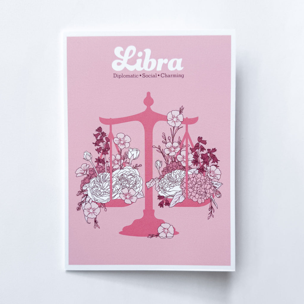 A Libra-themed greeting card featuring a pink background with a pink balance scale decorated with white and dark pink flowers, and the word 'Libra' printed at the top, shown against a white background.