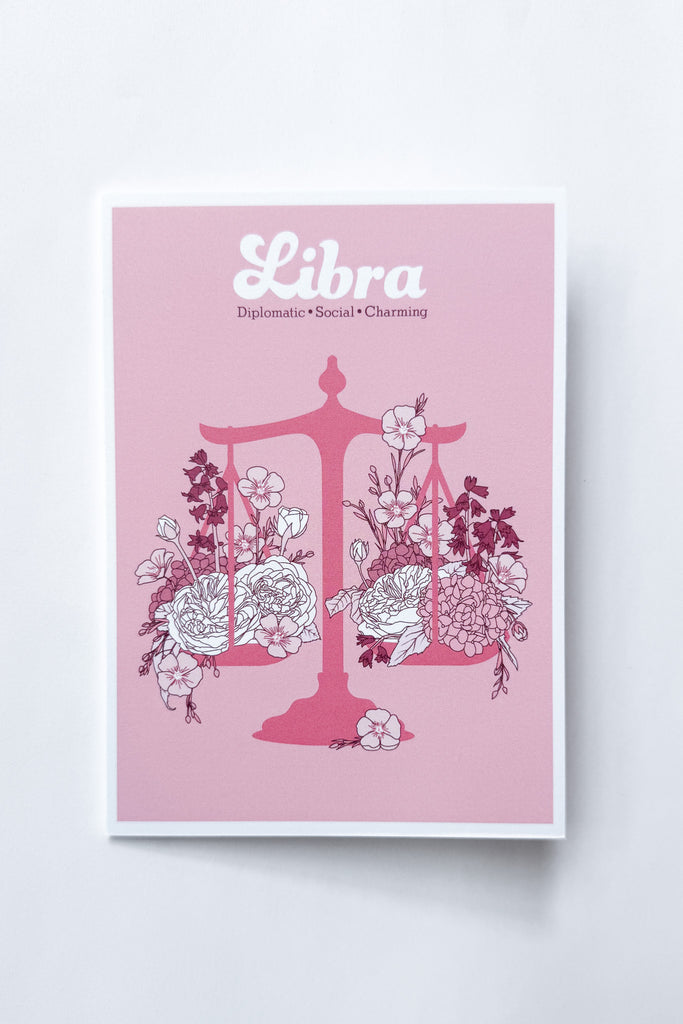 A Libra-themed greeting card featuring a pink background with a pink balance scale decorated with white and dark pink flowers, and the word 'Libra' printed at the top, shown against a white background.