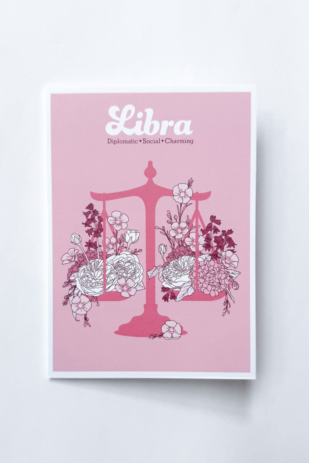 A Libra-themed greeting card featuring a pink background with a pink balance scale decorated with white and dark pink flowers, and the word 'Libra' printed at the top, shown against a white background.