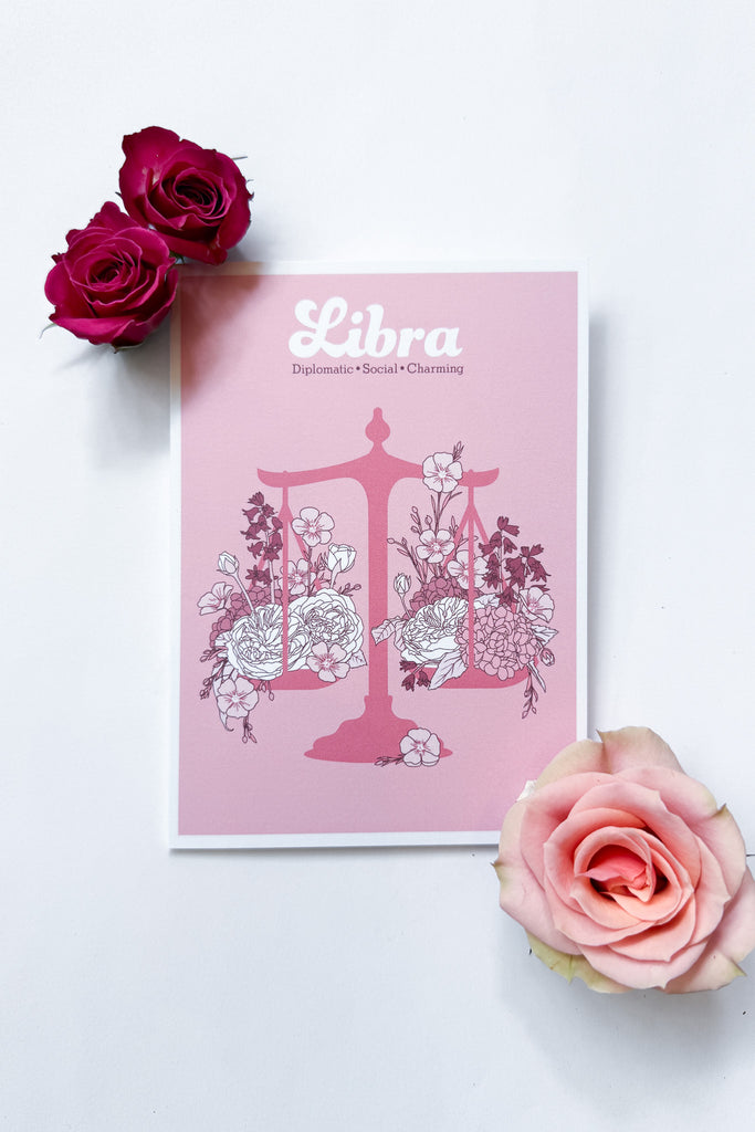 A Libra-themed greeting card featuring a pink background with a pink balance scale decorated with white and dark pink flowers, and the word 'Libra' printed at the top, shown against a white background with 3 roses.