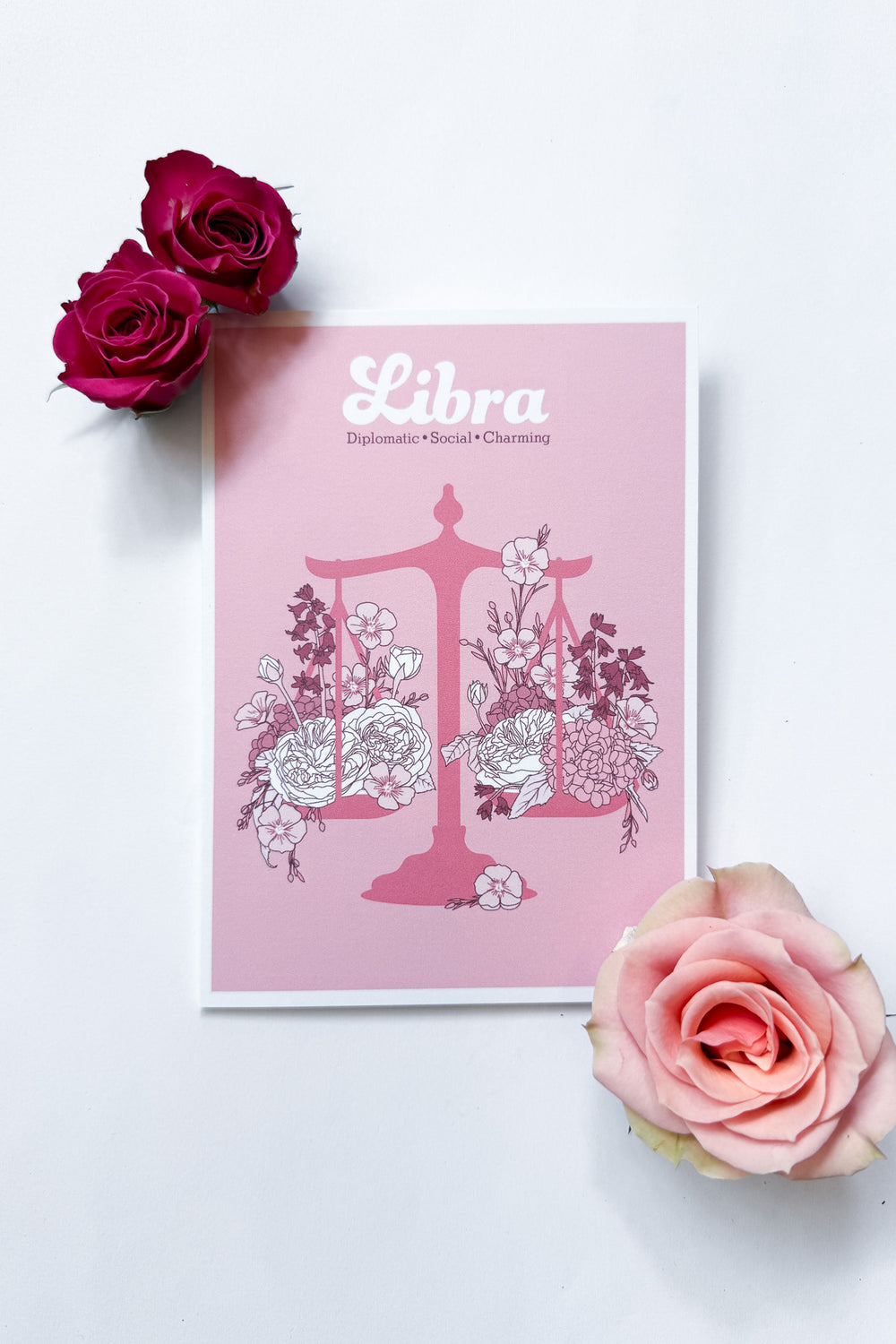 A Libra-themed greeting card featuring a pink background with a pink balance scale decorated with white and dark pink flowers, and the word 'Libra' printed at the top, shown against a white background with 3 roses.