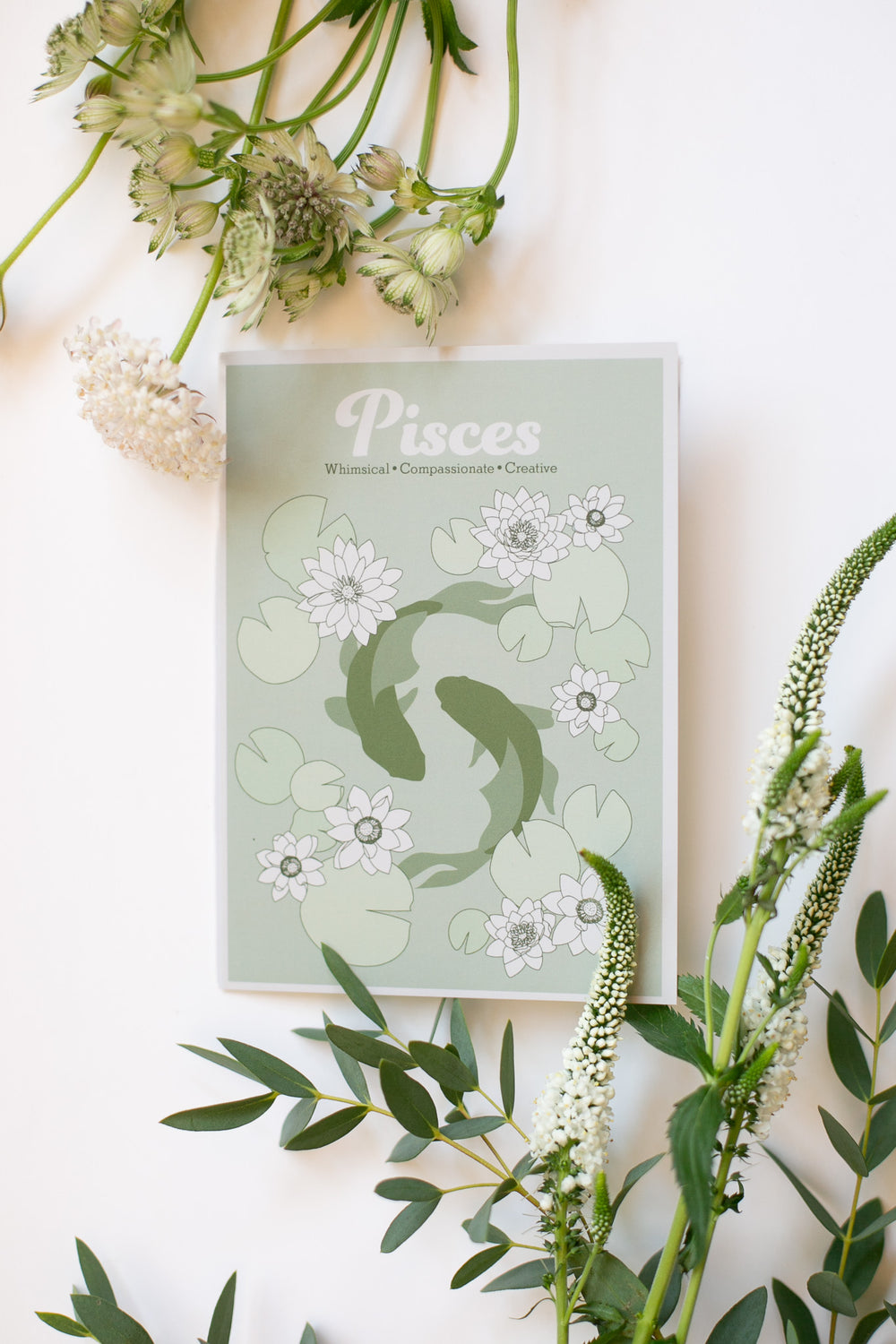 A greeting card titled ' Pisces' with a pale green background, white flowers, and two green koi fish, styled in a flat lay with green leaves and white flowers on a white surface.