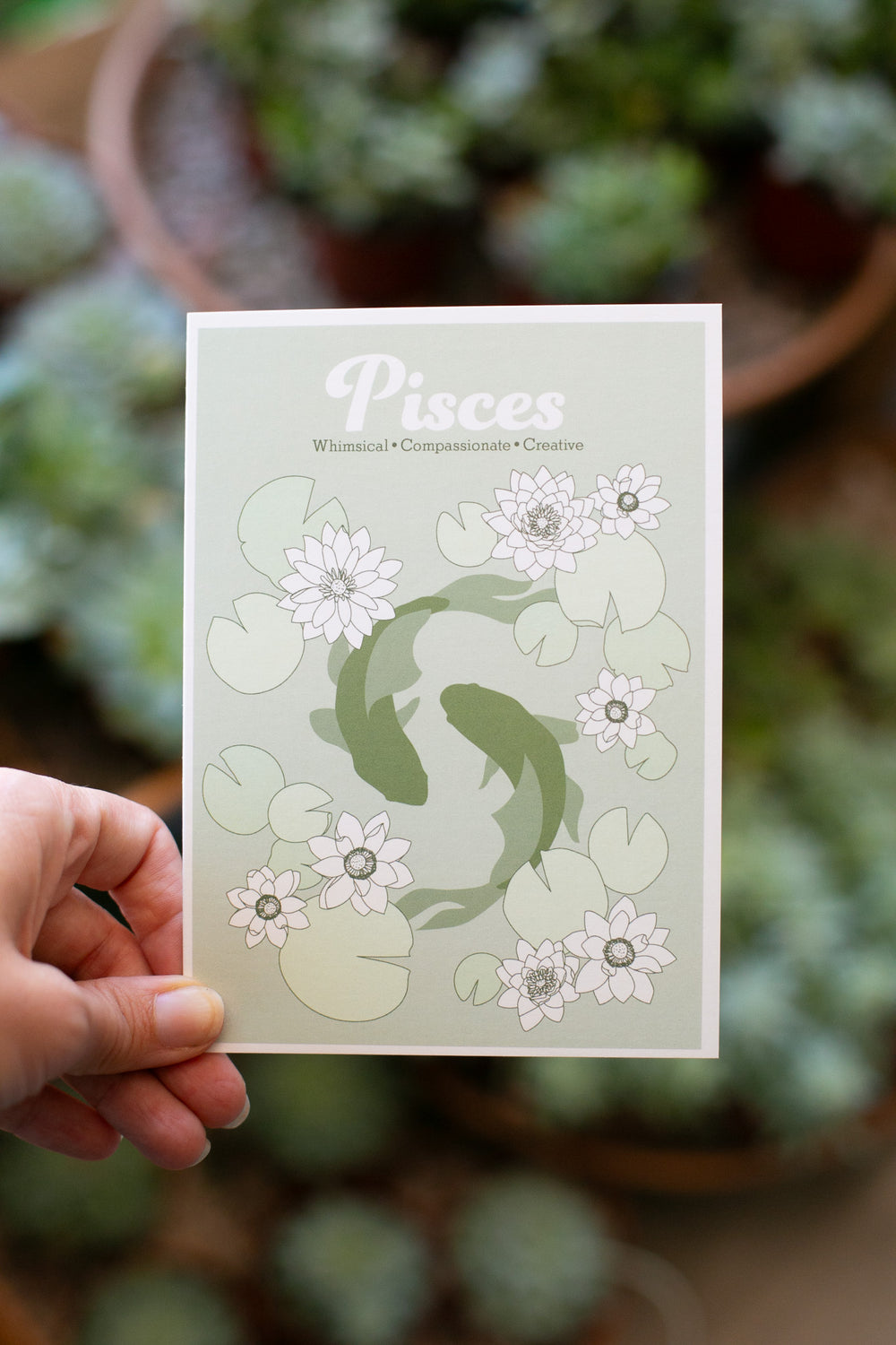 A hand holds a greeting card featuring a Pisces zodiac design with two koi fish, lily pads, and white flowers on a pale green background, with a blurred garden of succulents behind it.