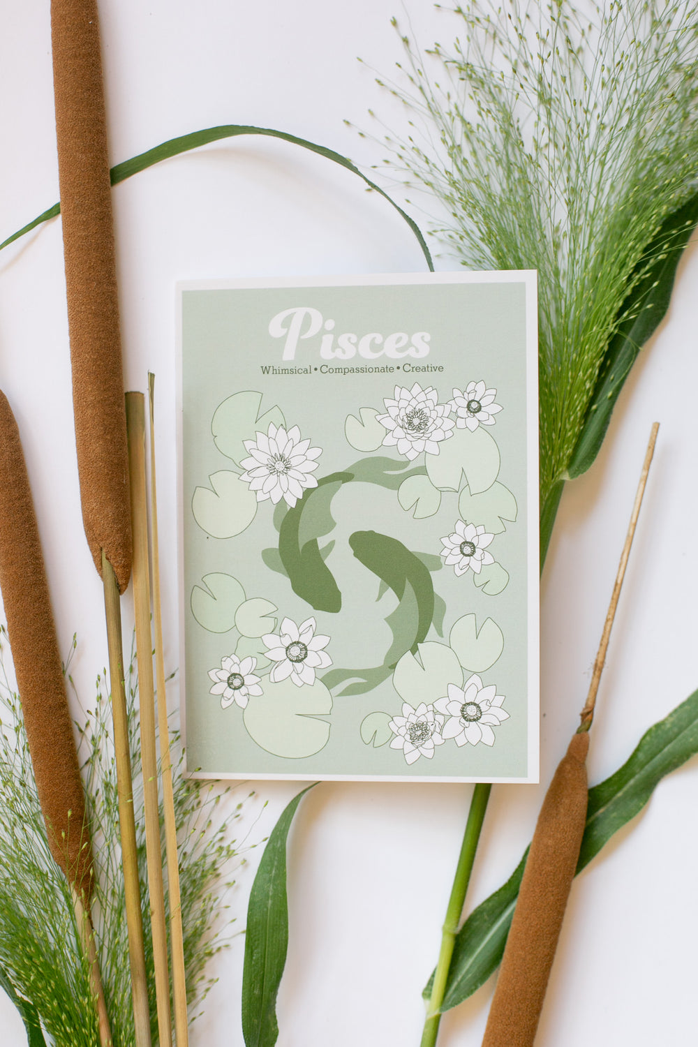 A Pisces zodiac-themed greeting card featuring two koi fish and white flowers on a pale green background, styled in a flat lay with cattails and green leaves on a white surface.