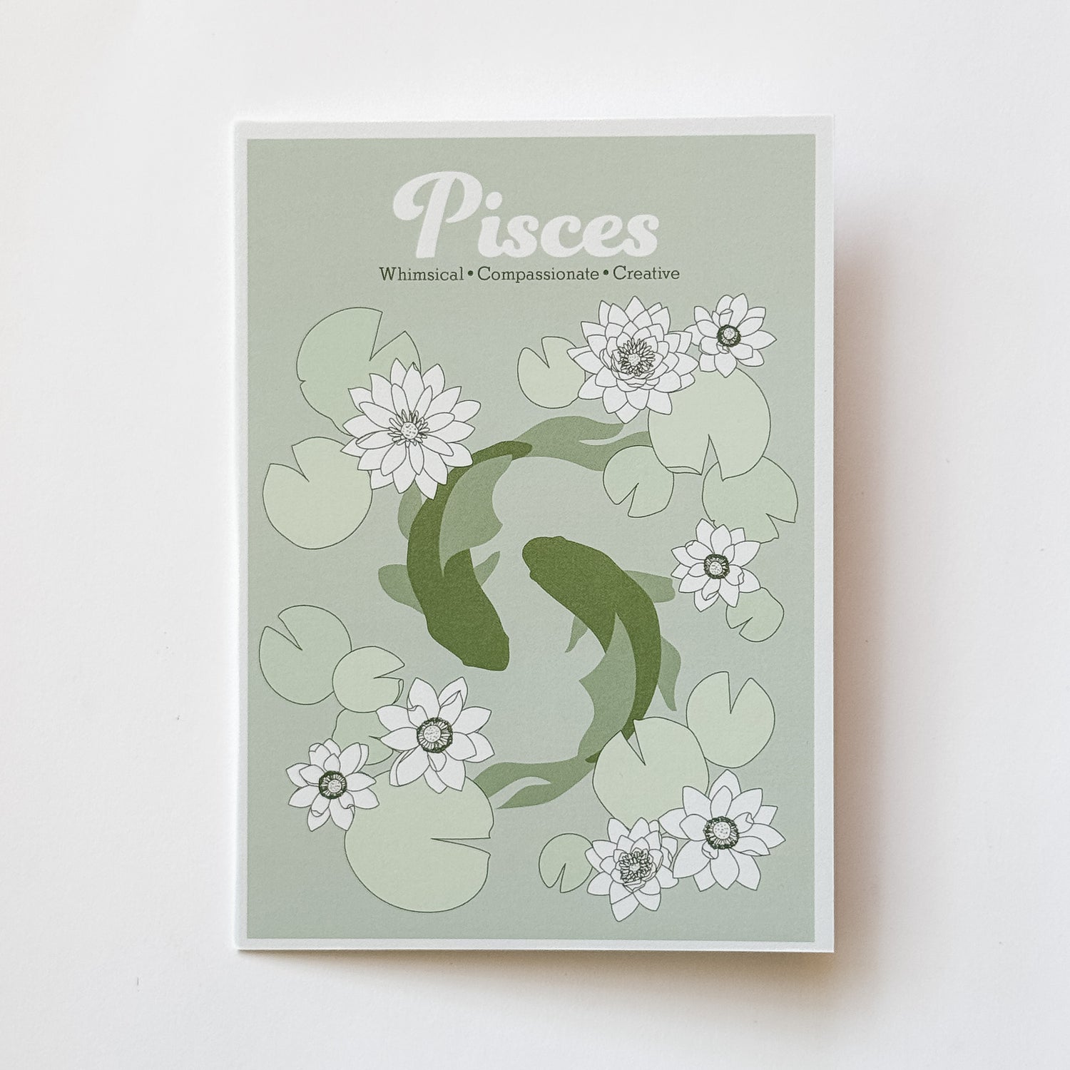 A greeting card featuring a Pisces zodiac design with two koi fish swimming among white lotus flowers and green lily pads on a pale sage background, photographed against a white background.