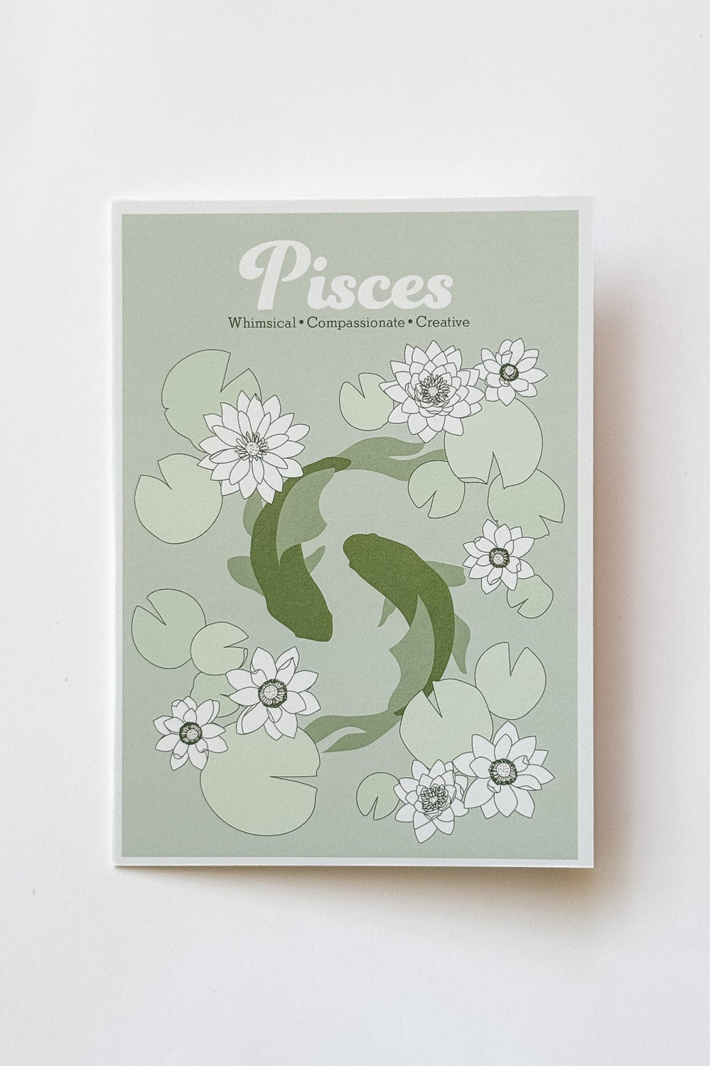 A greeting card featuring a Pisces zodiac design with two koi fish swimming among white lotus flowers and green lily pads on a pale sage background, photographed against a white background.