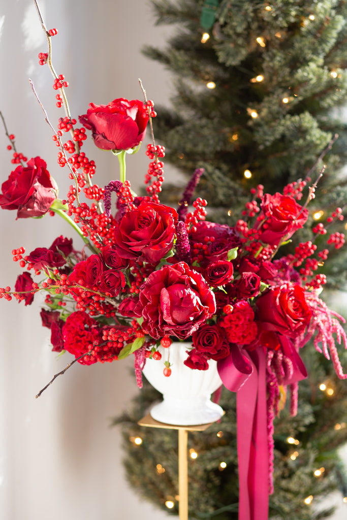 WildFlora's Red Velvet Arrangement in a white ceramic vase, wrapped with a fuchsia ribbon, in front of a Christmas Tree. It has red flowers, including rose, spray rose, berries, amaranthus, peony, and celosia.