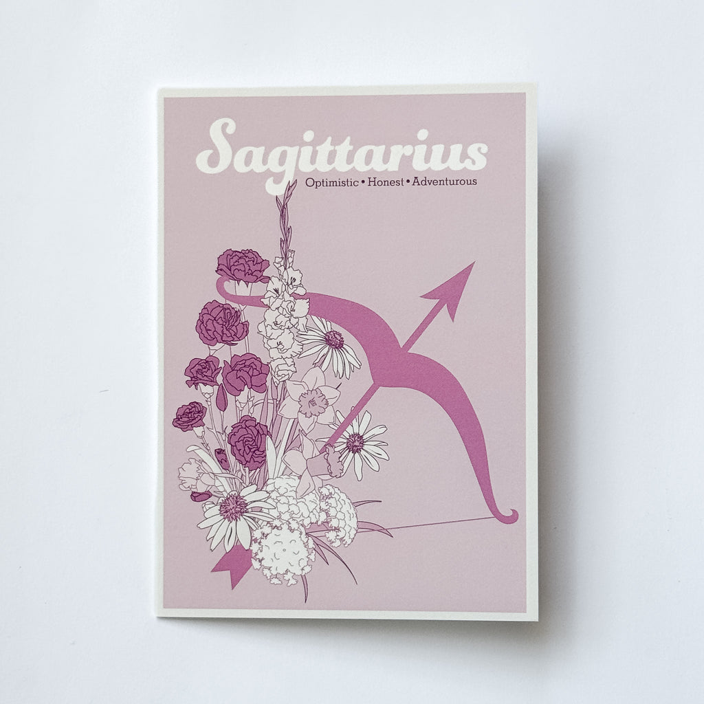 A greeting card featuring the Sagittarius zodiac sign, with a pink background, white and purple flowers, and a pink bow and arrow illustration, photographed against a white background.