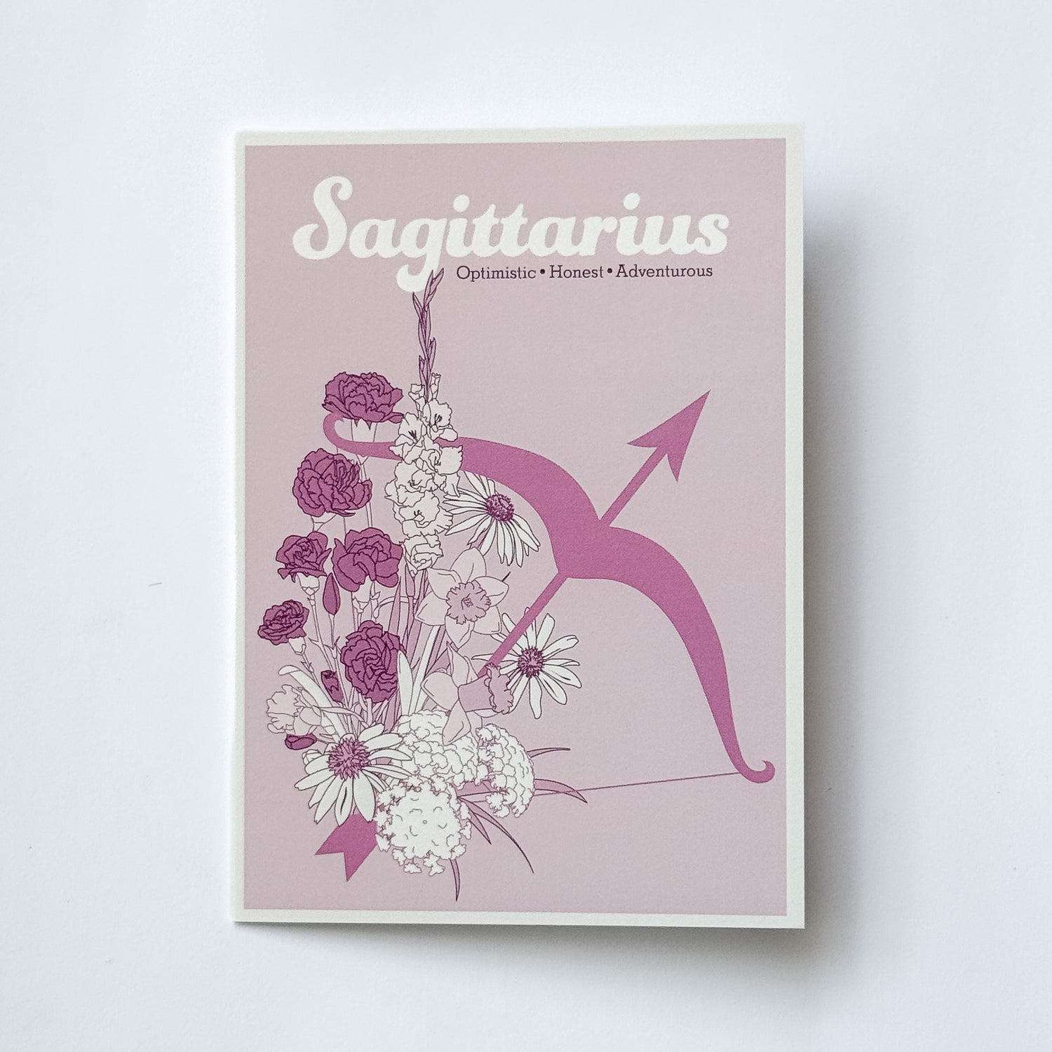 A greeting card featuring the Sagittarius zodiac sign, with a pink background, white and purple flowers, and a pink bow and arrow illustration, photographed against a white background.