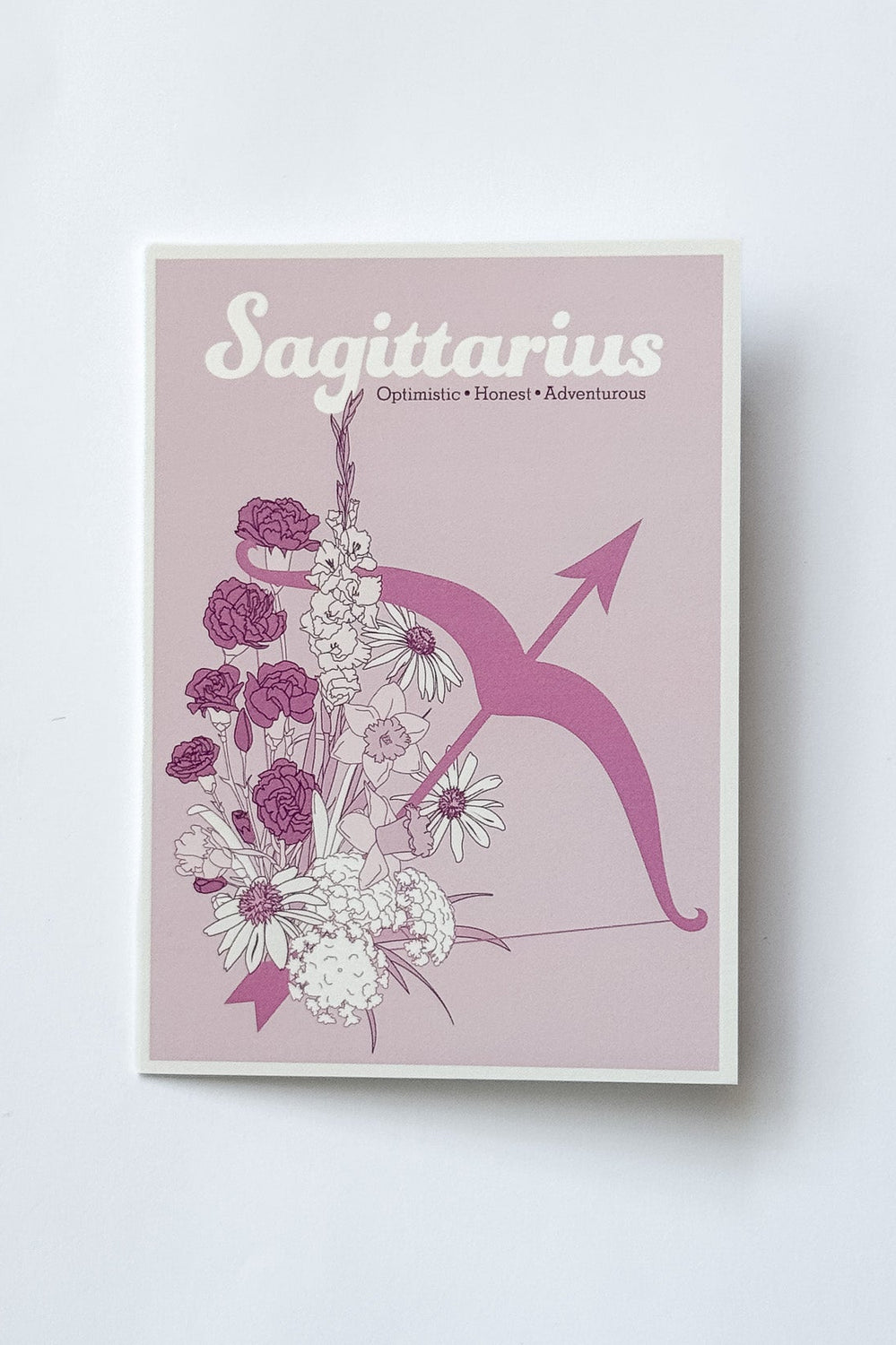 A rectangular card featuring the Sagittarius zodiac sign, with a pink background, white and purple flowers, and a pink bow and arrow illustration, photographed against a white background.