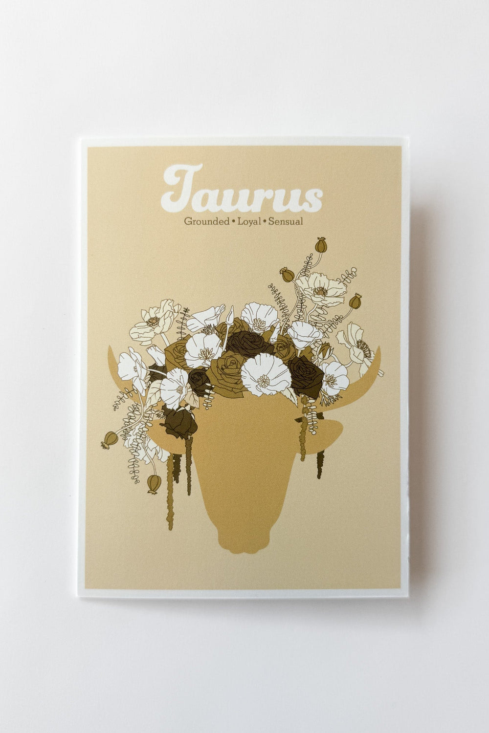 A greeting card featuring a beige background with the word 'Taurus' at the top and an illustrated silhouette of a bull's head filled with white and dark brown flowers. The print is photographed against a white background.