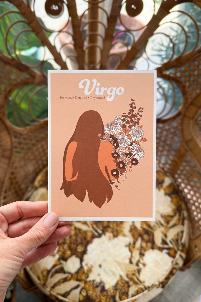 A hand holds a greeting card featuring a stylized brown silhouette of a woman with a floral bouquet on a peach background, with the word 'Virgo' at the top. The card is shown in front of a chair with a floral cushion.