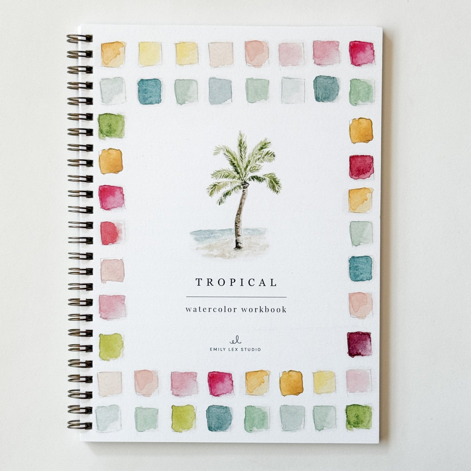 A spiral-bound watercolor workbook titled 'TROPICAL watercolor workbook' with a central palm tree illustration and a border of colorful watercolor swatches, photographed on a light neutral background.