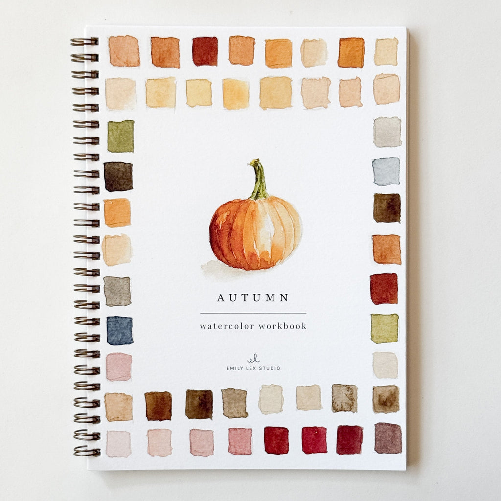 A spiral-bound watercolor workbook titled 'AUTUMN' with a central painted pumpkin and a border of watercolor color swatches in warm autumn tones, photographed on a light neutral background.