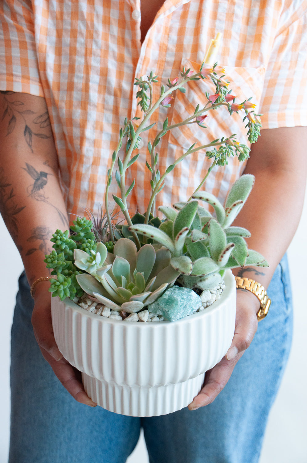 A woman in a white and orange checkered shirt holding a circular, ribbed white planter with succulents, air plants, and a crystal, sitting on a marble table in front of a white backdrop.