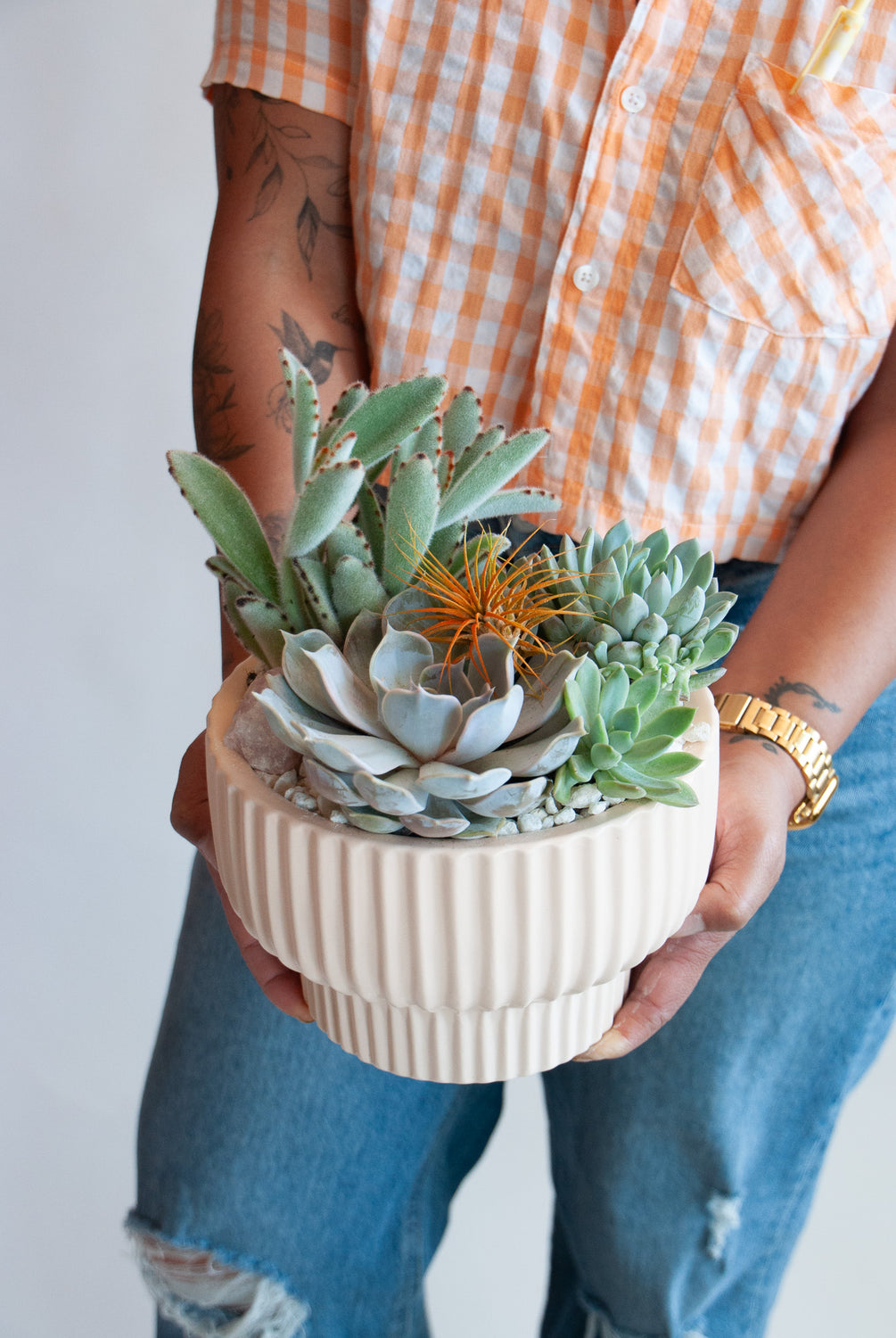 A woman in a blush-colored and orange checkered shirt holding a circular, ribbed white planter with succulents, air plants, and a crystal, sitting on a marble table in front of a white backdrop.