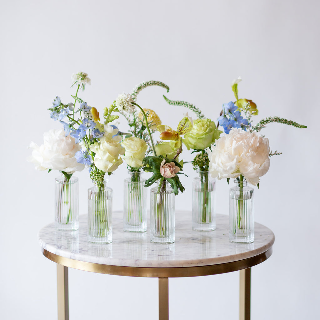 WildFlora's Holiday Sleigh Ride Bud Vases on a small marble table in front of a white backdrop. They have blue, green, white, and chartreuse flowers, and include rose, veronica, delphinium, scabiosa, hellebore, pitcher flower, and peony.