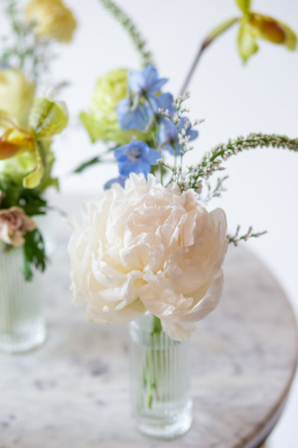 WildFlora's Holiday Sleigh Ride Bud Vases on a small marble table in front of a white backdrop. They have blue, green, white, and chartreuse flowers, and include rose, veronica, delphinium, scabiosa, hellebore, pitcher flower, and peony.