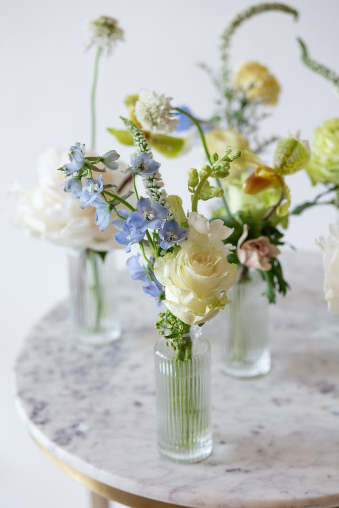 WildFlora's Holiday Sleigh Ride Bud Vases on a small marble table in front of a white backdrop. They have blue, green, white, and chartreuse flowers, and include rose, veronica, delphinium, scabiosa, hellebore, pitcher flower, and peony.