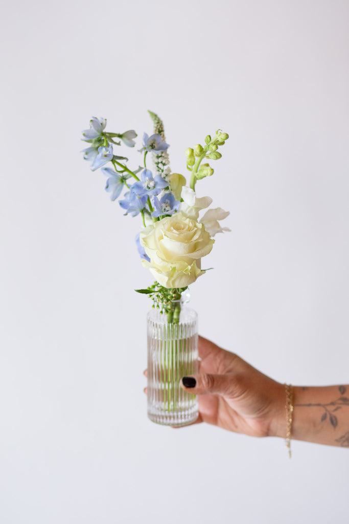 A hand holding one of WildFlora's Holiday Sleigh Ride Bud Vases in front of a white backdrop. They have blue, green, white, and chartreuse flowers, and include rose, veronica, delphinium, scabiosa, hellebore, pitcher flower, and peony.