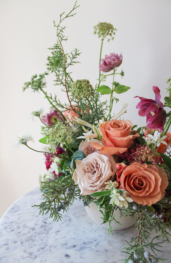 WildFlora's Winter's Garden Arrangement in a spherical grey vase, on a marble table in front of a white backdrop. It has sienna orange, pastel violet, red, pink, fuchsia, eggplant, and white flowers, and includes rose, scabiosa, hellebore, Queen Anne's lace, and juniper.