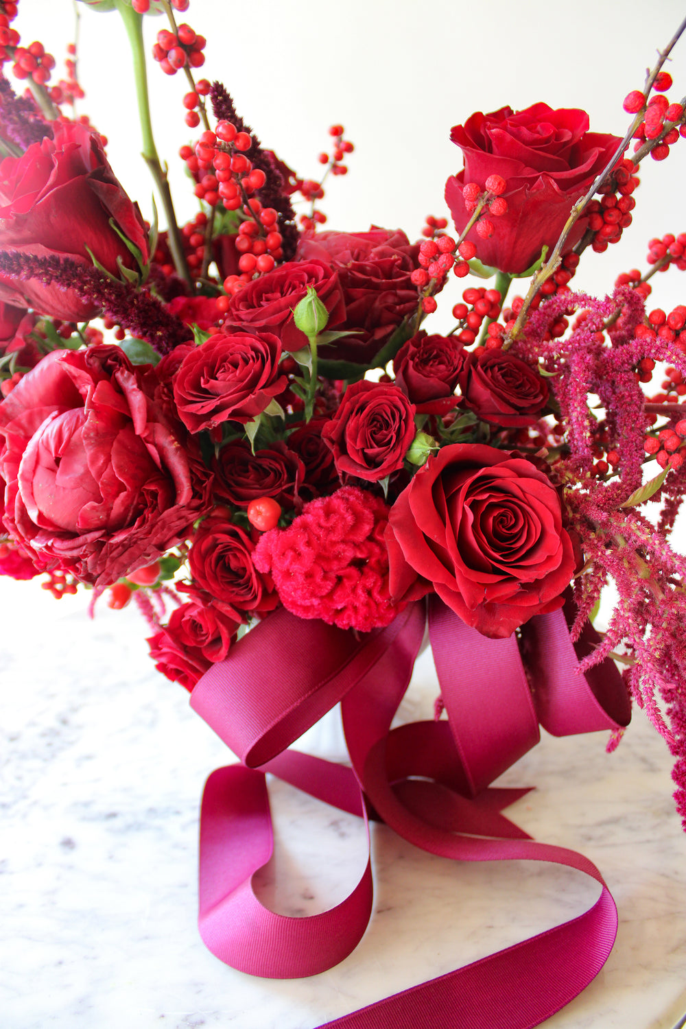 close up of WildFlora's Red Velvet Arrangement in a white ceramic vase, wrapped with a fuchsia ribbon, on a small marble table in front of a white backdrop. It has red flowers, including rose, spray rose, berries, amaranthus, peony, and celosia.